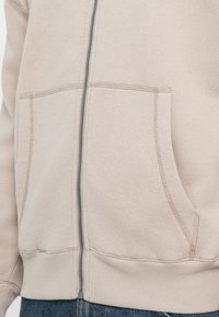 Hoodie bege com fecho éclair, feito de tecido suave, com dois bolsos frontais, detalhes cosidos e punhos e bainha canelados.