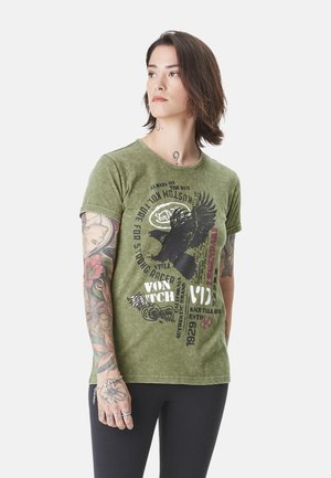Personne aux cheveux mi-longs portant un t-shirt graphique vert et un pantalon noir, montrant des tatouages floraux et calligraphiés sur son bras gauche.