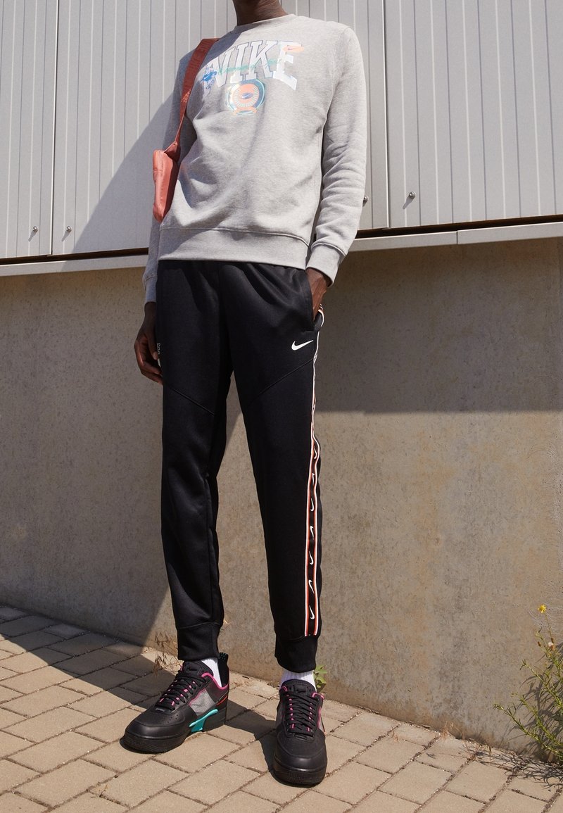 Person i grå Nike-tröja, svarta Nike-joggingbyxor med sidostråkar, svarta sneakers med rosa och turkosa detaljer, som står utomhus.