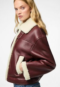 Giacca di pelle bordeaux con colletto e polsini in shearling crema, caratterizzata da una vestibilità rilassata e tasche laterali. Texture liscia e cuciture in vista.