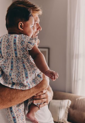 Muškarac drži bebu odjevenu u odjeću s uzorkom, oboje gledaju kroz prozor kojim ulazi svjetlo.
