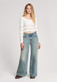 Maglione cropped bianco con righe orizzontali beige, abbinato a jeans in denim azzurro chiaro a gamba larga con finitura sbiadita e stile classico a cinque tasche.