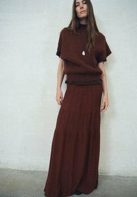 Femme se tenant contre un mur uni, portant un pull en tricot marron ample, une jupe plissée longue assortie et un collier avec un pendentif.