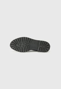 Parte inferior de una zapato negro que muestra un patrón de suela con ranuras y áreas elevadas, sobre un fondo blanco liso.