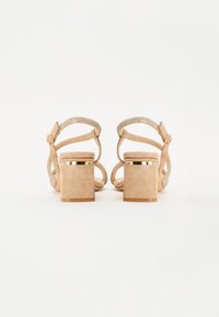 Sandales à talon carré en daim beige avec une bride métallique texturée, une boucle réglable à la cheville et une semelle plate lisse avec des accents dorés.
