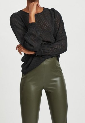 Personne portant un chemisier noir transparent avec un motif en zigzag et un pantalon en cuir taille haute vert olive, debout les bras croisés.