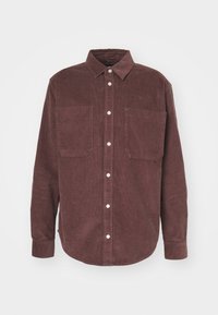 Chemise en velours côtelé marron à manches longues avec boutons sur le devant, col pointu et deux grandes poches poitrine sur fond uni.