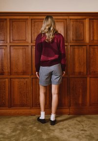 Άτομο με μακριά ξανθά μαλλιά, φορώντας μπορντό πουλόβερ, καρό σορτς, λευκές κάλτσες και μαύρα παπούτσια, στέκεται με την πλάτη σε ξύλινο τοίχο με πάνελ.