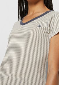 Femme portant un T-shirt rayé blanc et bleu marine en V avec un petit logo de baleine sur la poitrine gauche et des cheveux tressés.