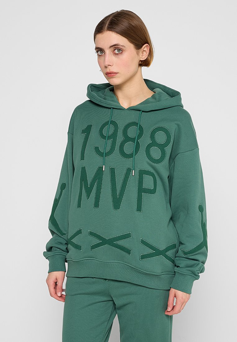 Jeune personne portant un sweat à capuche vert et un pantalon avec "1988 MVP" et des motifs de couture décoratifs, se tenant devant un fond clair et uni.