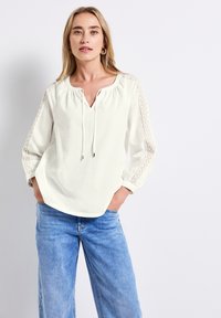 Vit blus med V-ringning, dragsnodd, och mönstrade genomskinliga ärmar. Kombinerad med ljusblå vida jeans. Mjuk tygtextur.