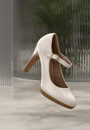 Beige Mary Jane high heel shoe with a gold buckle strap and wooden textured heel, positioned above a smooth gray floor.