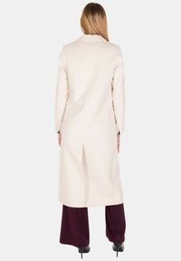 Cappotto di lana color crema con silhouette dritta, spacco singolo sul retro e maniche lunghe. Abbinato a pantaloni larghi bordeaux.