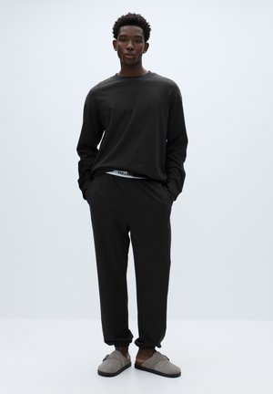 Czarna koszula z długim rękawem o luźnym kroju, połączona z pasującymi dresami z elastycznym paskiem w talii. Szare kapcie dopełniają strój.