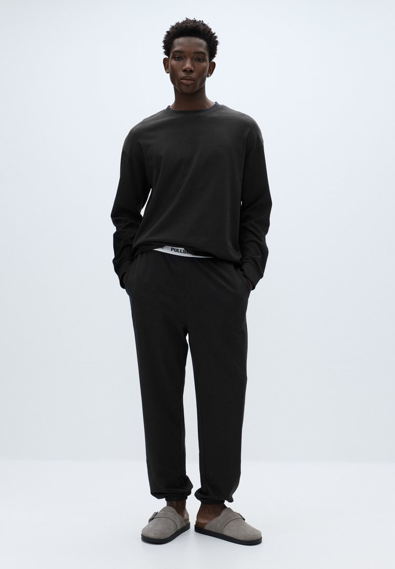 Černé tričko s dlouhým rukávem a volnějším střihem, spárované s odpovídajícími tepláky s elastickým pasem. Outfit doplňují šedé pantofle.