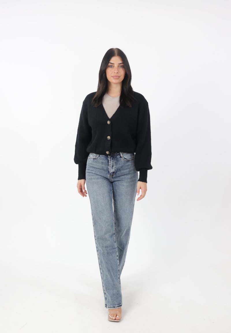 Schwarze Strickjacke mit V-Ausschnitt und drei großen Knöpfen, kombiniert mit hoch geschnittenen hellblauen Jeans und nudefarbenen Schuhen. Einheitlicher Hintergrund.