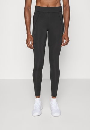 Leggings atletici neri con vita alta, caratterizzati da cuciture testurizzate e un design aderente, abbinati a scarpe da ginnastica bianche.