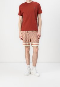 T-shirt en coton rouge avec un petit logo, associé à un short à motifs orange et beige, présentant des rayures horizontales et une ceinture élastique.