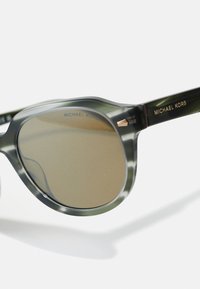 Krožne sončna očala s prosojnim zelenim okvirjem in rjavimi lečami. Imajo zlate dodatke in blagovno znamko "Michael Kors" na ročajih.