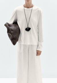 Witte blouse met lange mouwen, gecombineerd met lichtgekleurde wijde broek en een geweven donkerbruin tasje. Zwarte hangerketting voegt contrast toe.