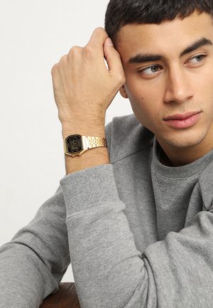 Gul digital klocka med rektangulär urtavla, armband i rostfritt stål, svart display och justerbar låsning, bärs på ett handled i en grå sweatshirt.
