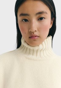 Pull à col roulé couleur crème avec une texture côtelée et un col haut, en tissu tricoté doux avec des détails de couture subtils.