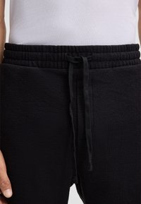 Zwarte sweatpants van een gestructureerde stof, met een elastische tailleband, trekkoord en zijzakken. Close-up zicht van de taille naar beneden.