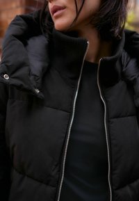 Giubbotto puffy nero con colletto alto, texture a coste e zip argentata. Presenta un design trapuntato e dettagli con bottoni a pressione.