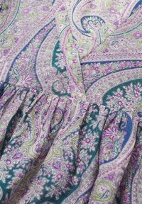 Tissu avec un motif paisley détaillé en tons de violet, vert, bleu et crème, présentant des dessins floraux et des volutes complexes sur un tissu doux et froncé.