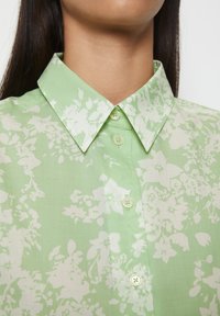Camicetta verde chiaro con un motivo floreale bianco, colletto a punte e chiusura con bottoni; realizzata in tessuto liscio con una consistenza morbida.