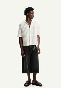 Camicetta bianca a maniche corte con un motivo a scacchi, abbinata a pantaloni shorts di denim neri oversize e sandali neri con dettagli a strappo.
