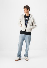 Sweat à capuche zippé gris clair avec une finition texturée, t-shirt graphique noir, jeans bleu clair et chaussures blanches avec des accents marron.