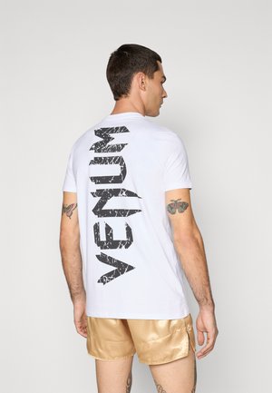 Hvid t-shirt med korte ærmer, der har et stort sort slidt "VENUM" logo trykt lodret på ryggen. Glat bomuldsstruktur.