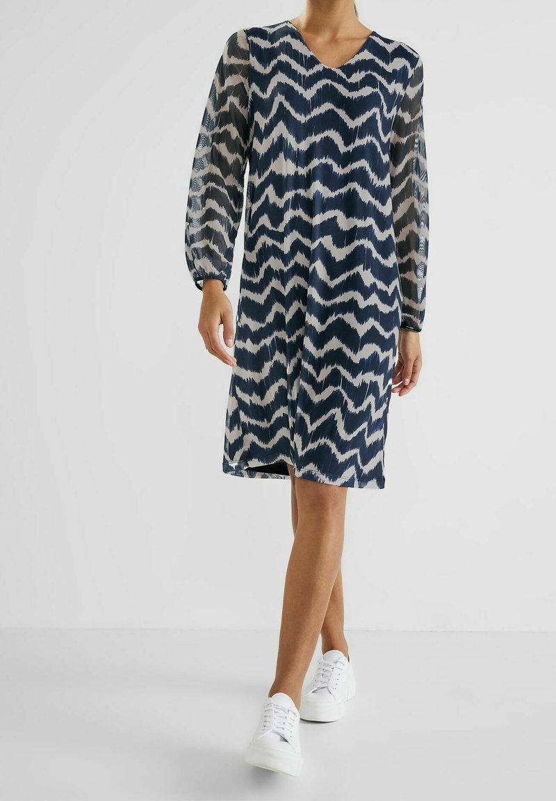 Femme portant une robe noire et blanche à motif zigzag jusqu'aux genoux, avec des manches longues transparentes et des baskets blanches, marchant vers l'avant.