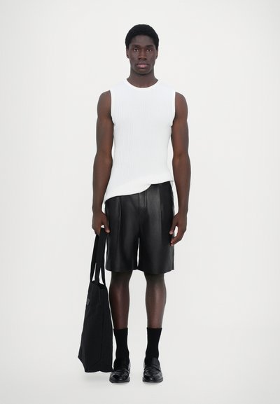 Haut sans manches blanc à côtes, shorts en cuir noir, sac noir avec poignées. Des mocassins noirs et des chaussettes complètent la tenue. Design minimaliste.