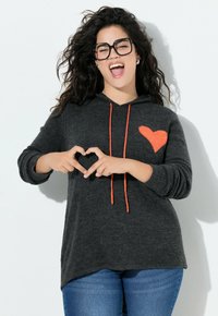 Sweat à capuche gris foncé avec un motif de cœur orange vif sur la poitrine, avec des cordons orange. Porté avec un jean bleu, modèle formant un cœur avec les mains.