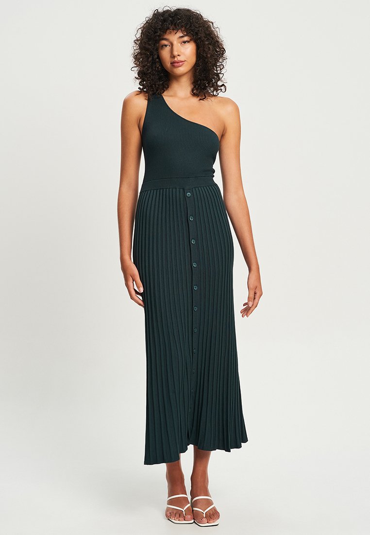 Dunkelgrünes, geripptes, einseitiges Kleid mit einem plissierten Rock und Knopfdetails auf der Vorderseite. Flache Schuhe ergänzen das Outfit.