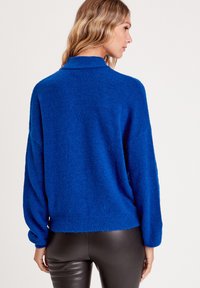 Blauer, fuzziger Pullover mit hohem Kragen, lockerer Passform und geripptem Saum. Hat lange Ärmel mit gerafften Bündchen. Kombiniert mit eleganten schwarzen Hosen.