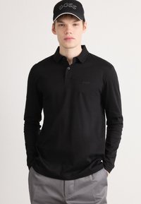Black long-sleeve polo shirt with a collared neckline and embroidered logo on the chest. The model also wears a black cap with a logo.