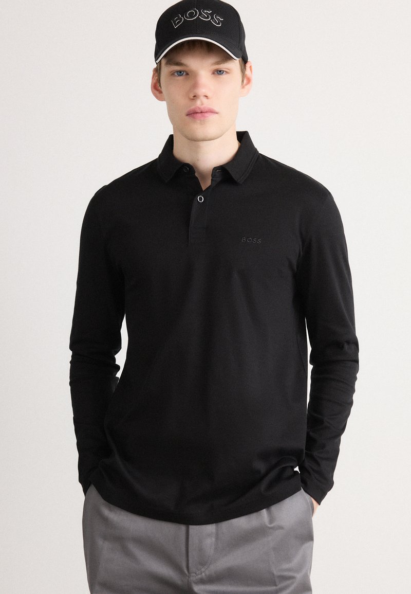 Black long-sleeve polo shirt with a collared neckline and embroidered logo on the chest. The model also wears a black cap with a logo.