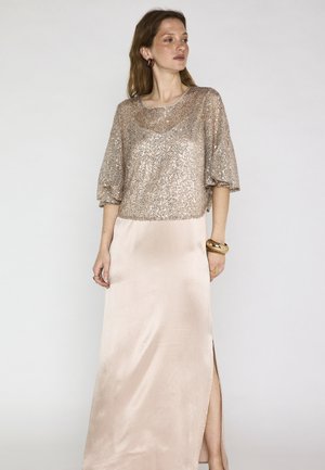 Femme portant un haut beige clair à sequins avec des manches jusqu'au coude et une jupe en satin assortie avec une fente sur le côté, accessoirisée avec des bijoux en or.