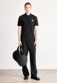 Melna īsroku polo krekls ar zeltainu emblēmu kreisajā krūtīs, kopā ar melnām biksēm un apaļu melnu somu ar divām rokturiem.