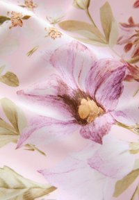 Blommönstrad tyg med en rosa bakgrund som har stora lila och krämfärgade blommor, ömtåliga gröna blad och en mjuk, slät textur.