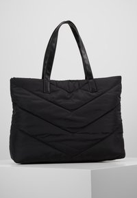 Sac en toile matelassé noir avec couture en chevron et poignées noires lisses, posé sur une surface blanche contre un fond gris.
