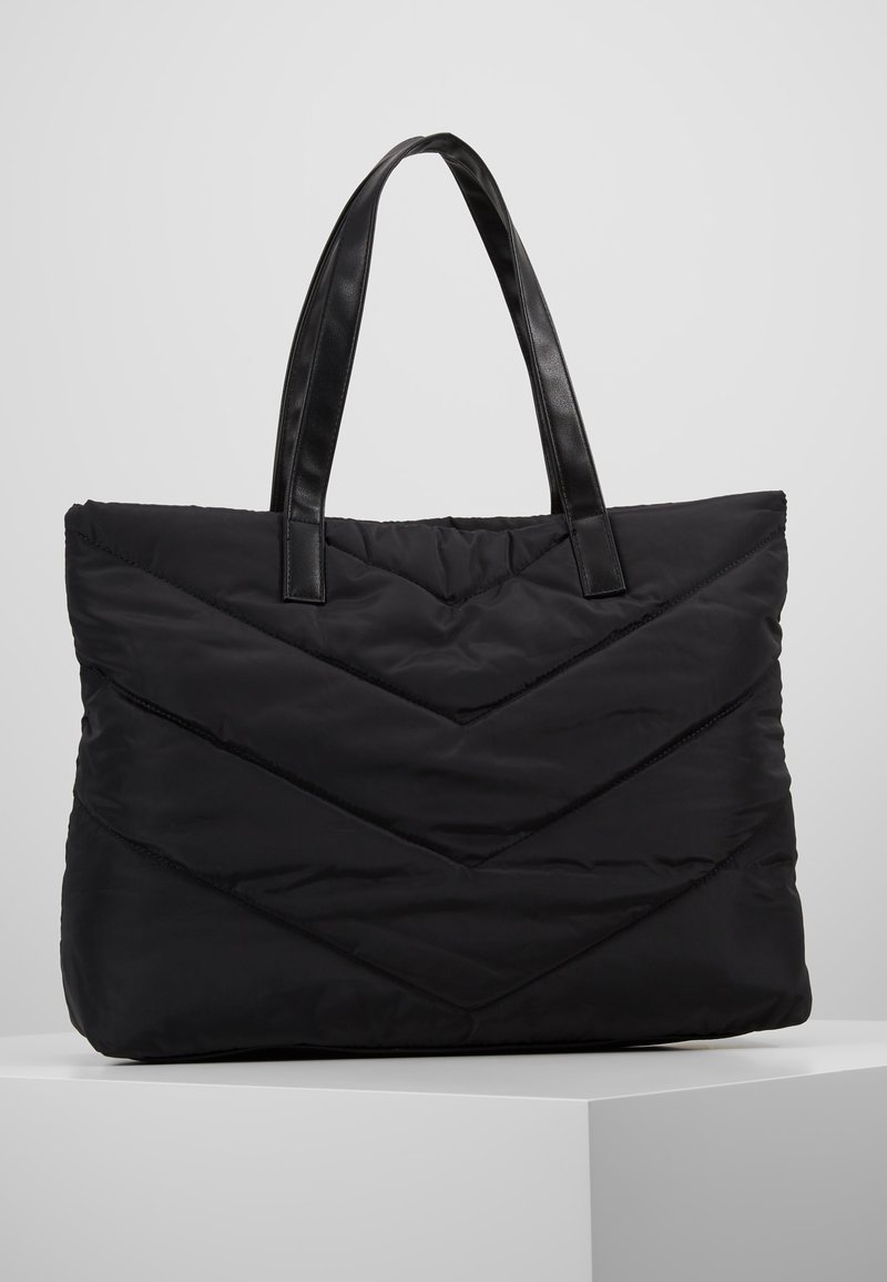 Sac en toile matelassé noir avec couture en chevron et poignées noires lisses, posé sur une surface blanche contre un fond gris.