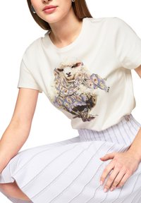 White t-shirt featuring an illustrated sheep wearing patterned fabric. The model wears a textured white skirt with vertical stripes.