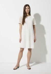 Weißes Spitzenkleid mit kurzen Puffärmeln, das mit floralen Ausschnitten und einem lockeren Schnitt versehen ist, kombiniert mit schwarz-weißen Riemchensandalen.