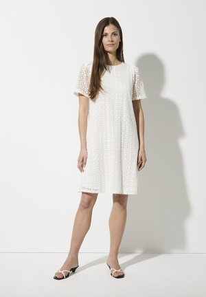 Weißes Spitzenkleid mit kurzen Puffärmeln, das mit floralen Ausschnitten und einem lockeren Schnitt versehen ist, kombiniert mit schwarz-weißen Riemchensandalen.