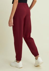 Persona che indossa pantaloni jogger bordeaux e sneakers bianche, in piedi con una gamba leggermente sollevata su uno sfondo neutro.