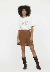 Shorts en velours côtelé marron à taille haute et plis sur le devant, associés à un t-shirt blanc ample avec un design imprimé, et des bottes marron au genou.